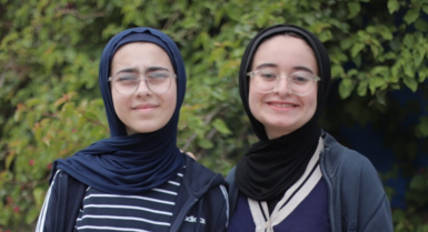 Tala and Farah Mousa, two teeanged girls. Both have their heads covered in hijabs, wear glasses and navy zip-up jackets, and are smiling at the camera in front of a large green tree.