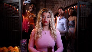 A young woman with bleached blonde shoulder length hair wears a pink top and holds a drink in a glass with straw held in two hands at waist level. Behind her a group of friends takes a selfie in a dimly lit bar or nightclub.ford, who plays Anna Knight