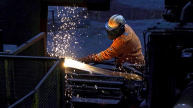 A steelworker wearing orange protective gear welds as sparks flies against a dark background in Scunthorpe Steel works in 2025.