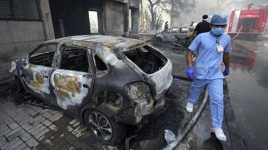 A man in medical scrubs walks beside a burnt out car