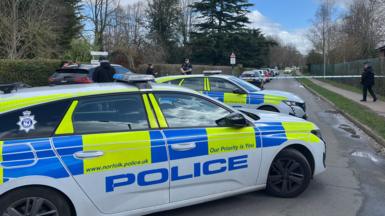 Two marked fluorescent yellow, blue and white police car. Blue and white police tape has been put across the road to cordon it off. There are police officers at the cordon.