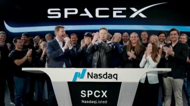 Elon Musk wearing a black leather jacket, stands in the center of crowds and in front of a SpaceX sign.