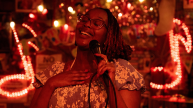 Edna Nicole Luckett sings at a microphone, her hand on her heart, with light-up signs showing musical notes behind her