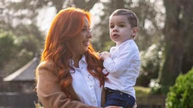 A woman with red hair, wearing a white blouse and brown jacket, smiles at a little boy who she is holding in her arms. He is wearing a white shirt and jeans and has short dark hair.