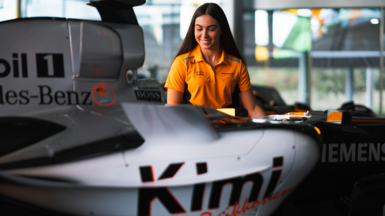 Ella is sitting next to a McLaren F1 car while she is wearing an orange, team-branded T-shirt 