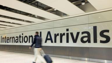 An airport corridor with the words 'International Arrivals' printed in large black letters onto a white wall. A man can be seen walking in front of the sign with a wheeled suitcase, beige trousers and a dark jacket, but he is slightly blurred to indicate the fact he is moving quickly. 