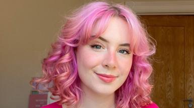 A woman with wavy pink hair in a pink top smiles at the camera for a selfie, taken indoors