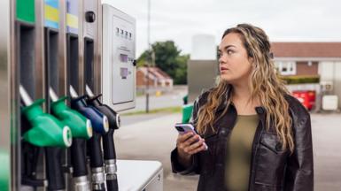 A young woman stands at the petrol pumps in a petrol station and looks at the prices
