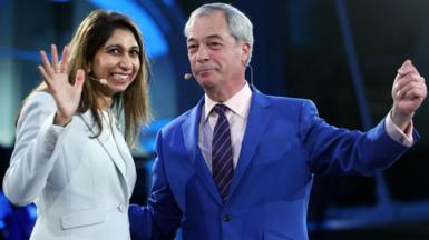 Suella Braverman waves with her right hand as she grins and moves towards Nigel Farage who smirks as he holds aloft his left arm. Both are wearing blue suits and have small headset microphones.