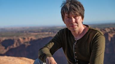 Prof Brian Cox sits on a hillside. He is wearing a green top and is staring directly at the camera.