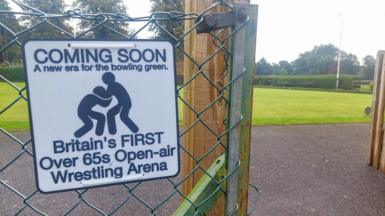 A joke sign reads: Coming soon. A new era for the bowling green. Britain's first over 65s open-air Wrestling Arena