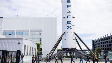 A row of people walk past a large white rocket with blue "space x" logo written on it.
