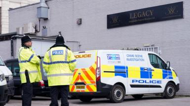 Two police officers wearing yellow jackets and black helmets stand outside a white bricked building. A crime scene investigation police van is parked underneath a black sign that reads Legacy Independent Funeral Directors