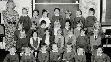 A black and white photo of Gwen Mayor, a blonde-haired woman in a patterned dress, and her 28 pupils arranged in four rows of seven in a classroom. The children are in school uniform.
