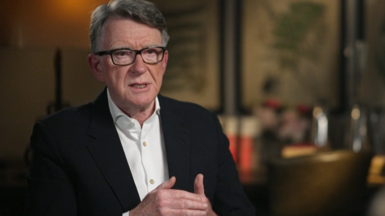 Lord Mandelson sits in a chair while being interviewed. He is wearing a white shirt and a black blazer. He is also wearing glasses and has greying dark hair, styled in a side parting. Also visible is the blurry background of what looks to be a modern looking room.