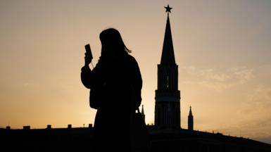A person is silhouetted against a glowing orange sky as they look at their smartphone, with the silhouette of the Kremlin in the background during sunset in the Red Square in Moscow on 31 March.