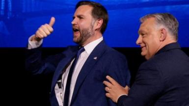 JD Vance and Orban stand together smiling, in front of their national flags