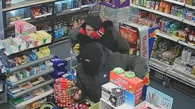 A still from CCTV footage showing two men standing at the counter of a convenience shop. One is wearing a black jacket and a black balaclava, while pointing an imitation hand gun. The other is wearing a black hooded jacket with a red scarf covering his face.