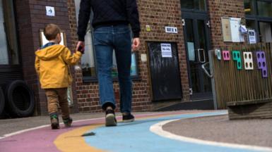 A small boy wearing a yellow raincoat holds his dad's hand as they walk into a polling station in a primary school.
