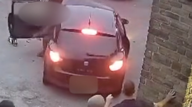A black with bright red reverse lights on. The passenger-side door is being thrown open. A group of people watch from behind