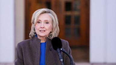 Hillary Clinton in a purple suit and brown coat, standing in front of a black microphone