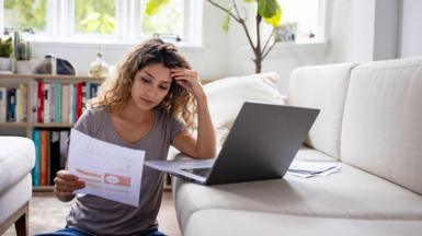 A woman has her hand pressed against her head as she sits on the floor with her laptop on the sofa looking at her energy bill on a piece of A4 paper