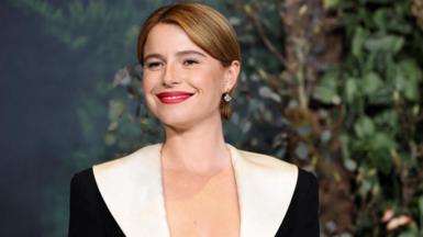 Jessie Buckley smiles at the camera. She has light brown hair, and is wearing a black and white dress