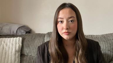 Laura is a young woman with long, brown hair. She is staring at the camera. She is sitting on a grey sofa and is wearing a brown pinstripe blazer and a black t-shirt.