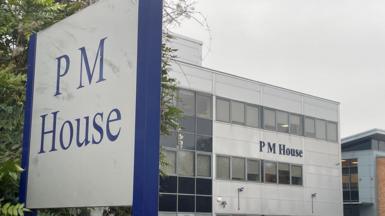 A large office building with the name “PM House” displayed prominently on its exterior. The building has a modern, rectangular design with multiple rows of windows and light grey cladding. In the foreground, a tall sign also reads “PM House” in blue lettering on a white background, framed by a dark blue border. Several cars are parked in front of the building, suggesting a business or office complex. 