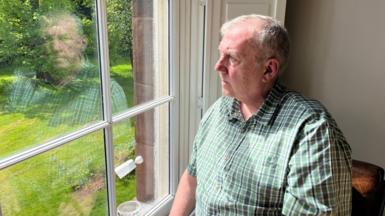 Alan Jones, 59, who has thinning grey hair and is wearing a green chequered short-sleeved shirt, sits by a first-floor window looking out onto a green garden lined with trees. 