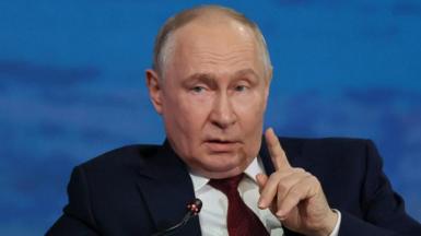 President Putin lifts his finger as if to make a point as he speaks into a microphone. He is dressed in a navy blue suit, white shirt and red patterned tie and is speaking in front of a marine blue backdrop