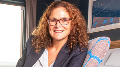 Jo Boydell sat on a chair, smiling and looking at the camera. She wears glasses and has shoulder-length curly light brown hair.