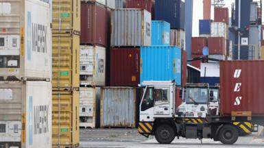 A white truck in front of stacks of different-coloured cargo containers at a port
