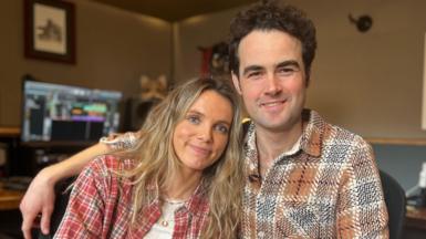 A woman with blonde hair and a red checked shirt leans into her husband who has his arm around her. He has a brown checked shirt and black hair. 