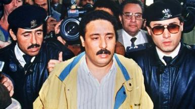 Fhimah (centre) being escorted by security officers and surrounded by journalists before a hearing in 1992 in connection with the Lockerbie bombing. He has short brown hair and a moustache and is wearing a yellow jacket and a white and blue striped short.
