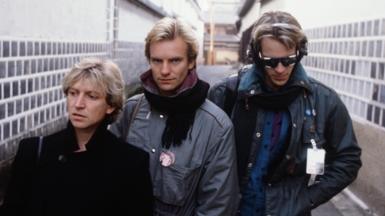 Andy Summers, Sting and Stewart Copeland in an alleyway in Japan in 1981 - they are wearing winter clothing, jackets and scarfs. Sting is the only one looking at the camera
