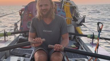 Man with long hair sits in a rowing boat, behind him is equipment, dusk is falling over the Pacific Ocean