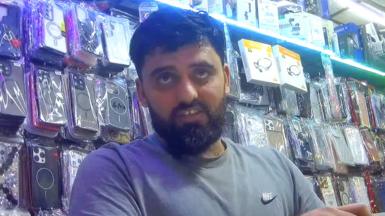 Shop worker inside a mobile phone accessories shop, behind a glass counter, with walls densely covered in packaged phone cases, headphones and electronic accessories.