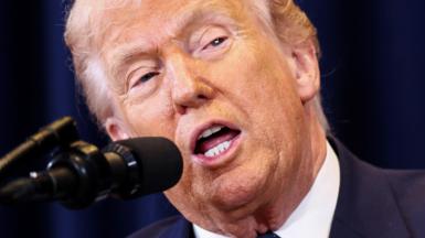 US President Donald Trump speaks into a microphone, his mouth is agape and he is wearing a blue suit jacket and white shirt. Behind him is a dark blue curtain.