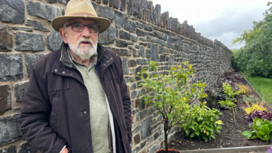 John Watkeys standing against his new stone garden wall which is about head height. He is wearing a fawn-coloured hat and glasses with a black coat and has a white beard. A flower bed can be seen to the right of the wall and grass to the right of that. 