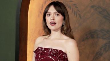 Aimee Lou Wood, who has long brown hair and is wearing a strapless burgundy dress, poses at a The White Lotus premiere in Los Angeles