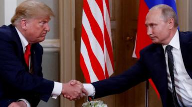 Trump on the left and Putin on the right shake hands. They two are not smiling but rather pursing their lips and narrowing their eyes. The Russian and US flags are behind them. 