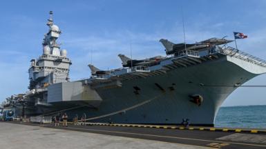 The aircraft carrier pictured in 2025 while moored up in Singapore during a media tour, against a blue sky with partial cloud cover.