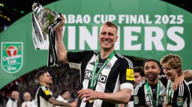 A player in a black and white Newcastle United shirt holds a silver trophy aloft with one hand as his team-mates celebrate around him. In the background an arched green banner reading 'Carabao Cup Final 2025 winners' is visible.