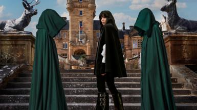 Claudia Winkleman stands on the castle steps, flanked by two hooded figures in green cloaks.