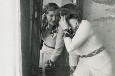 Posed black and white photo of Zo looking into a mirror while wearing a white dress with decorative cuffs and belt