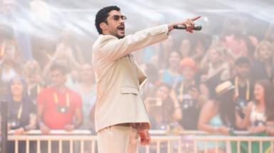 Bad Bunny in a beige suit points at the crowd with a mic in his hand. The audience are faded in the background.