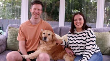 A man and woman sit on a grey couch, with a golden retriever dog between them. The man wears an orange t-shirt and black short. The woman wears blue jeans and a black and white striped jumper. Behind them are clear windows with white frames. Outside are green trees and a black fence, separating the trees from their garden 