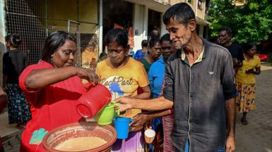 A woman pouring tea from a pot into the cup held by a man. Behind them is a queue of people waiting to get tea.