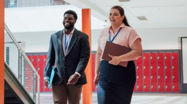 Two employees walk together while smiling. They're both wearing lanyards and holding folders. 